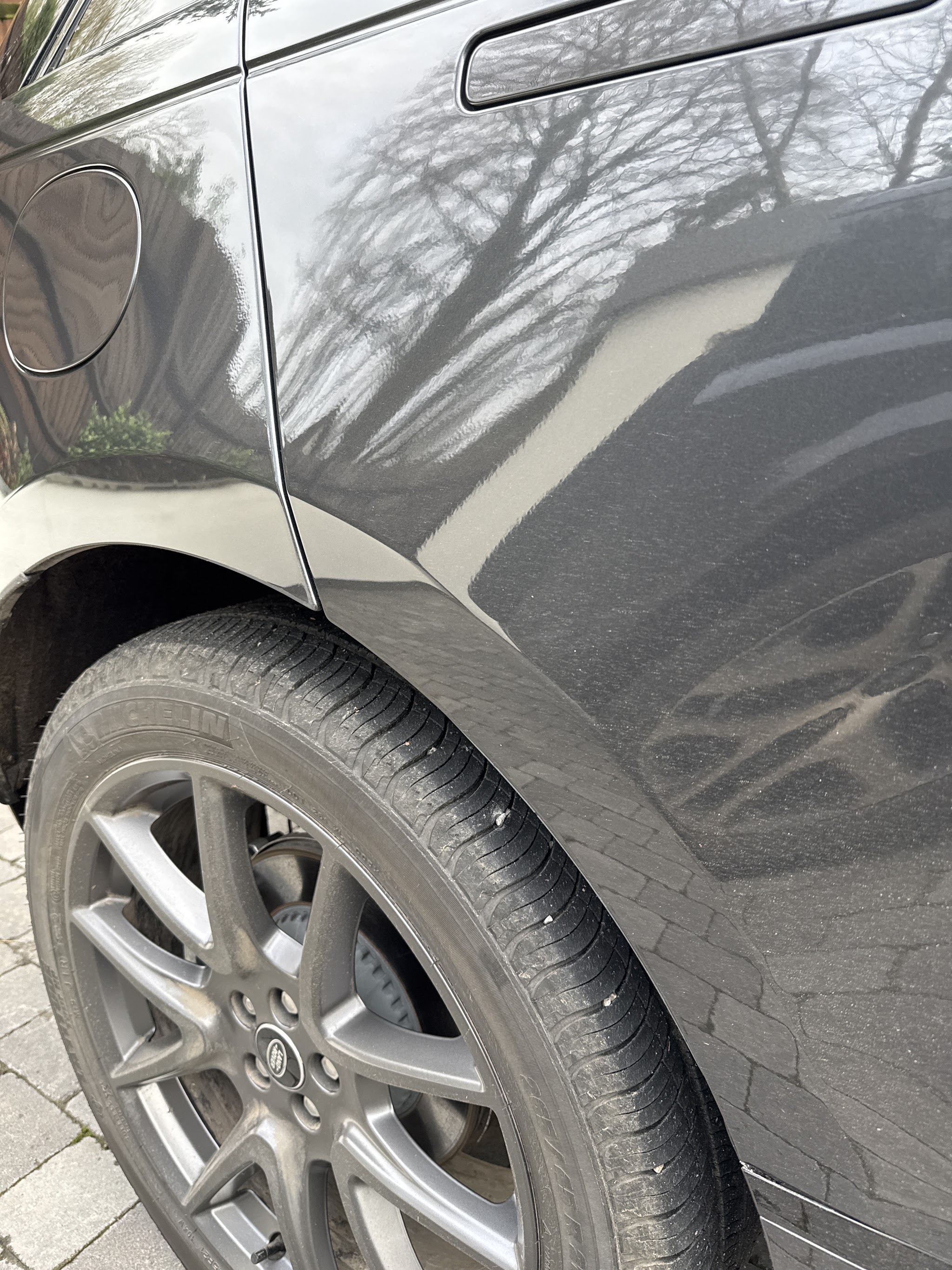 Trees reflected in the glossy dark grey paint of a car’s rear wing above the wheel